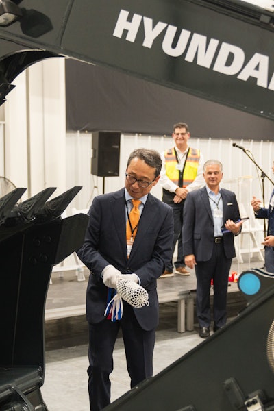 President and CEO of HD Hyundai XiteSolution, Young Cheul Cho, inaugurates the opening of the HD Hyundai North American Customization Center with the celebratory breaking of a bottle of wine over the bucket of one of the Hyundai excavators customized at the new facility.
