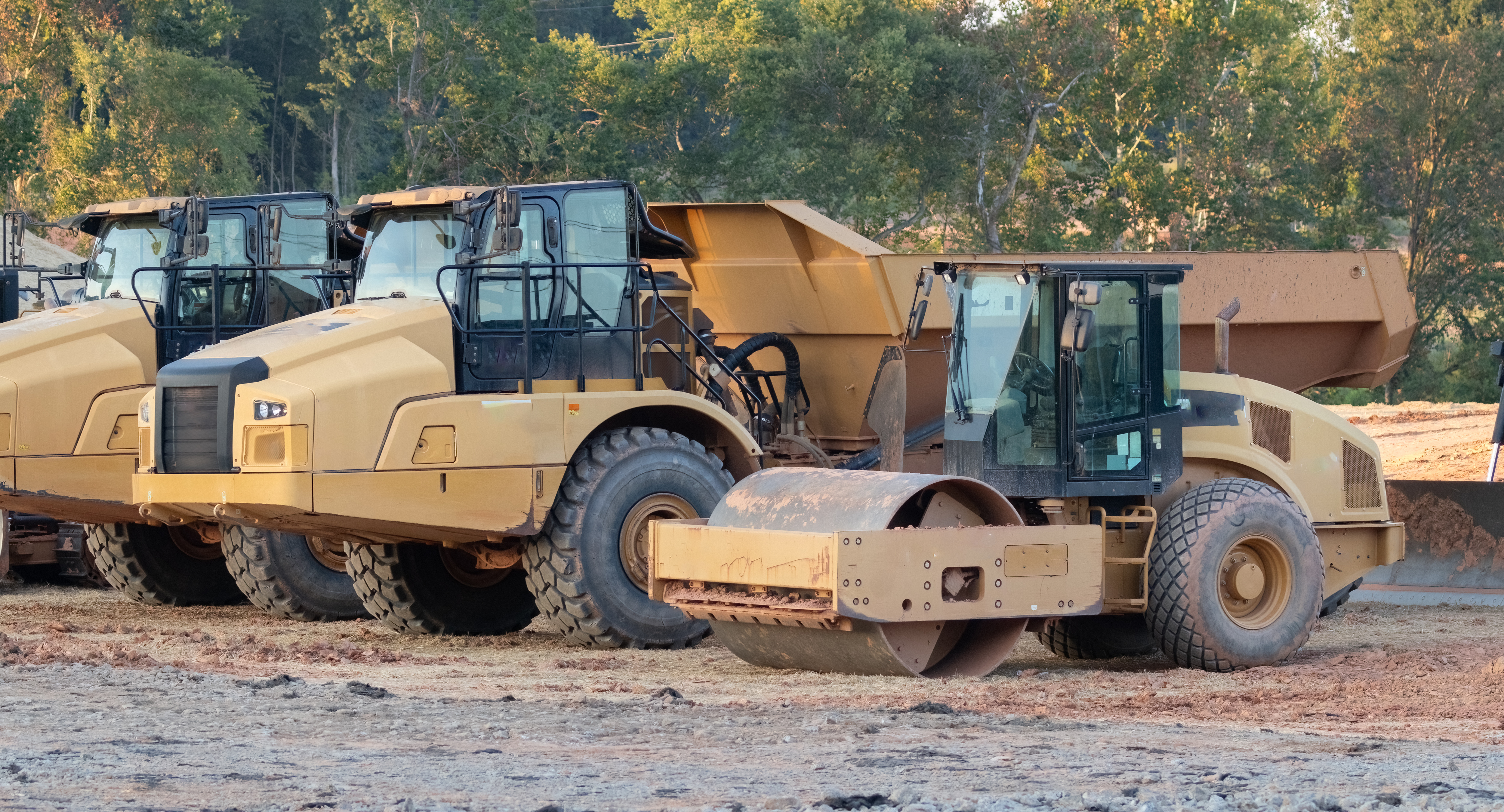 Heavy construction equipment including tan compactor and asphalt roller parked at job site with autumn trees in background