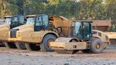 Heavy construction equipment including tan compactor and asphalt roller parked at job site with autumn trees in background