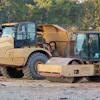 Heavy construction equipment including tan compactor and asphalt roller parked at job site with autumn trees in background