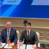 Two businessmen sign documents at formal meeting with Rexroth and Kawasaki logos displayed on screen behind them