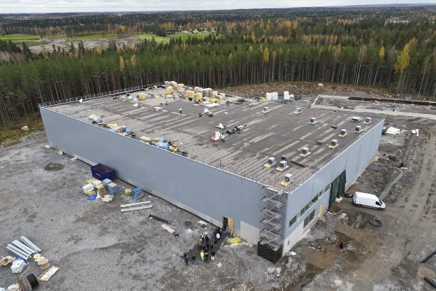 Aerial view of large white warehouse building under construction surrounded by forest with materials on roof