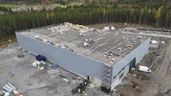 Aerial view of large white warehouse building under construction surrounded by forest with materials on roof