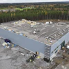 Aerial view of large white warehouse building under construction surrounded by forest with materials on roof